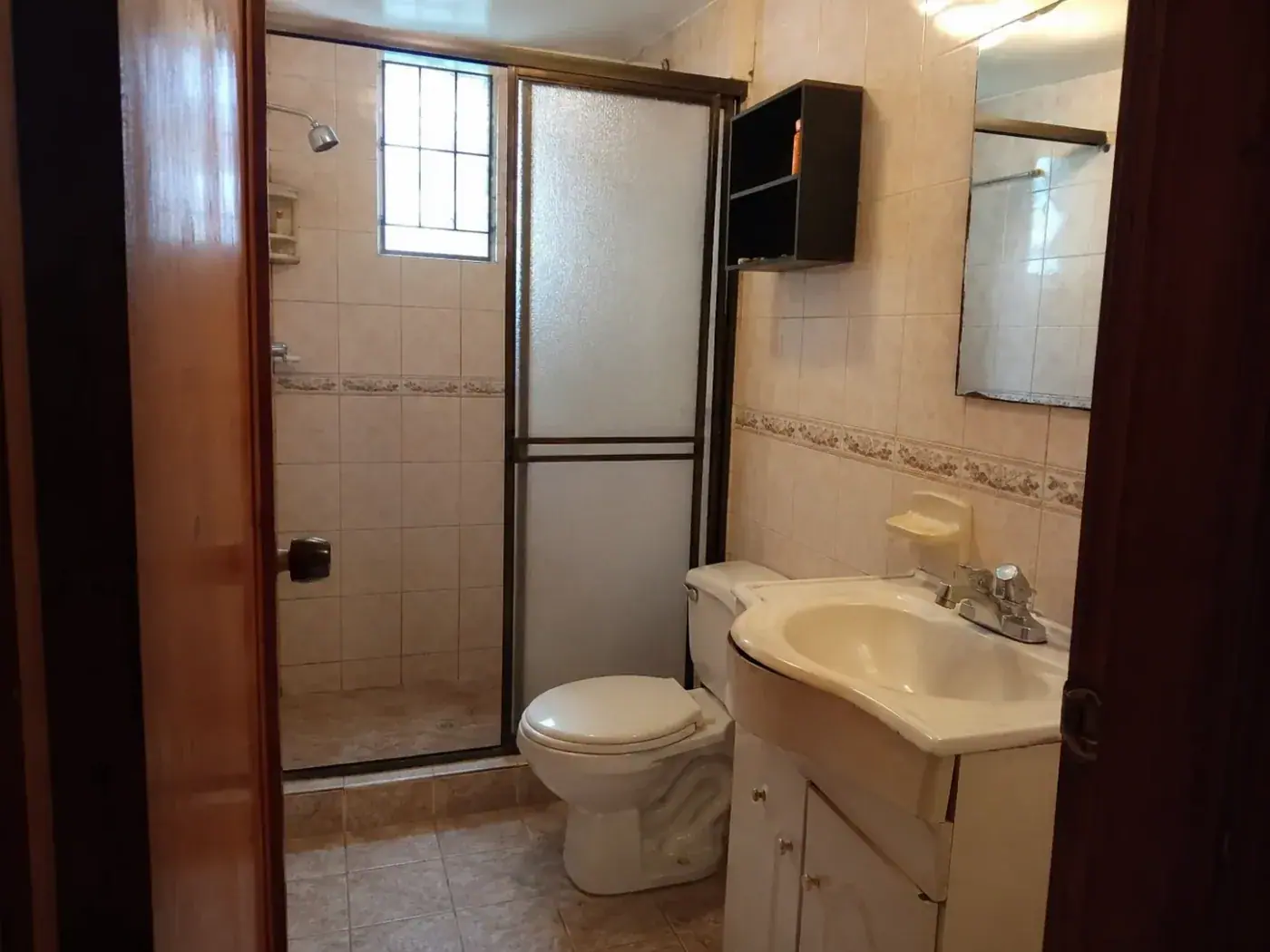 Baño antes de la remodelación: enchape beige con cenefa floral, ducha con puerta corrediza de vidrio corrugado y estante negro sobre el sanitario