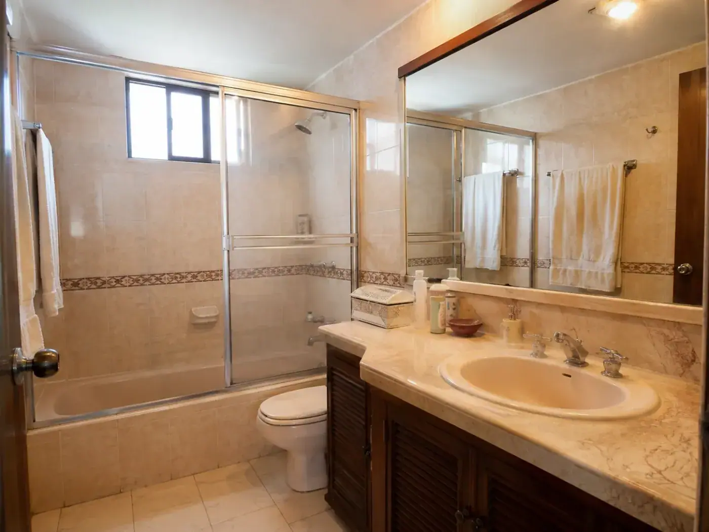 Baño antes de la remodelación: enchape beige con cenefa decorativa, puertas corredizas de aluminio sobre la tina y mueble de madera calada